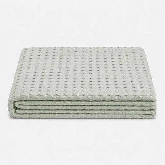 Couverture rafraîchissante Nuvia en fibre de bambou pliée, avec texture gaufrée respirante et coloris vert sauge doux sur fond clair.